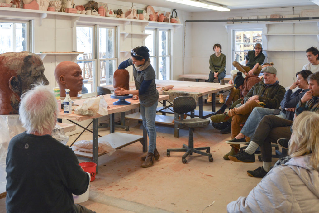 Cristina Cordova sculpting a head from red paper clay while a ring of students looks on