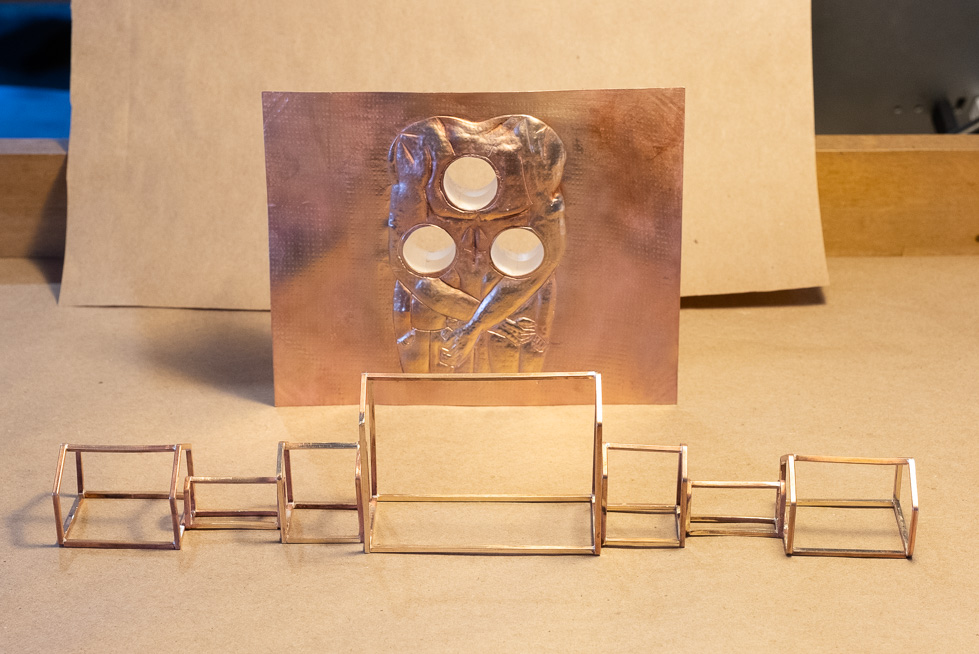 A sheet of copper with a relief image and three holes sits in front of an architectural model made from small copper bars