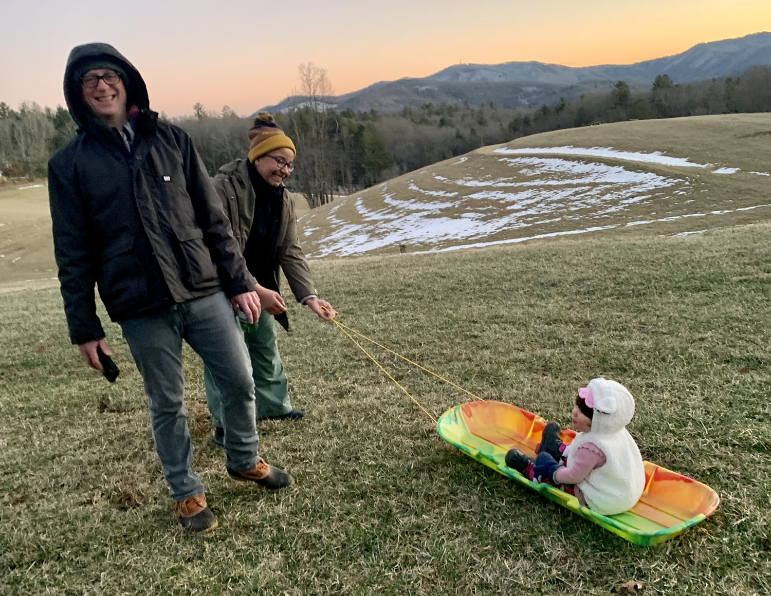 A man and a woman in a grassy field in winter, pulling a sled with a baby sitting in it
