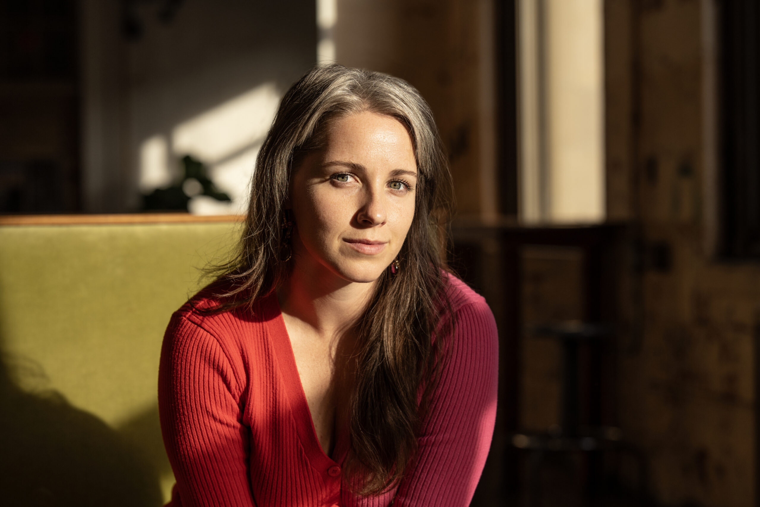 Portrait of Sarah Rachel Brown. Sarah has long brown hair, is wearing a red sweater, and is lit with natural light from a nearby window.